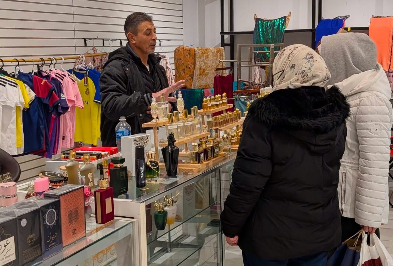 Man behind glass counter displays perfumes to two women in winter coats at a store with clothes and accessories.